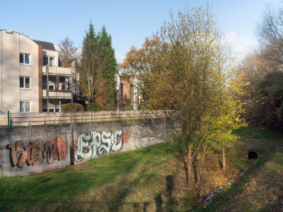 Mauer mit großflächigem Graffiti englang eines Baches, Häuser hinter der Mauer