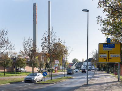 Eine verkehrsreiche Straße mit Weg-Hinweisschildern, zwei Industrieschornsteine im Hintergrund