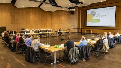 Circa 30 Menschen in einem Sitzungssaal an einer U-förmigen Tischformation, schauen auf eine Beamer-Präsentation