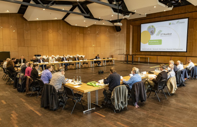 Circa 30 Menschen in einem Sitzungssaal an einer U-förmigen Tischformation, schauen auf eine Beamer-Präsentation