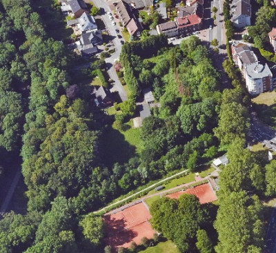Luftaufnahme eines Stadtgebiets mit Häusern, Bäumen, einem Asche-Sportplatz und Fußwegen